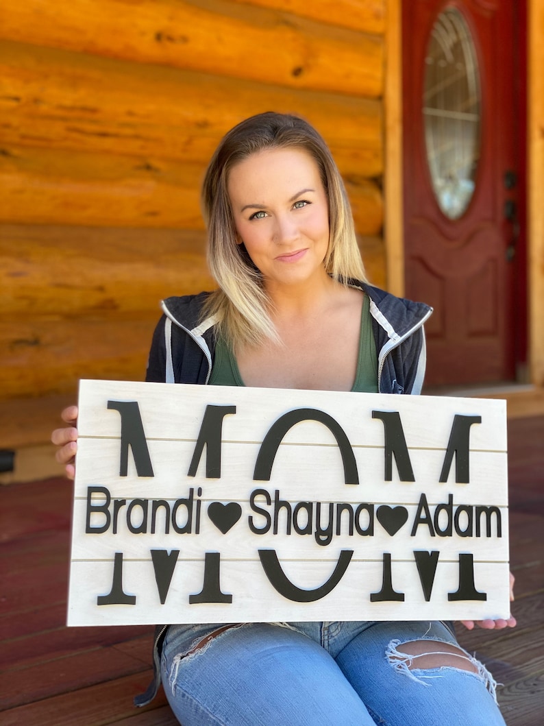 May include: A white wooden sign with the word "MOM" in large black letters. Below the word "MOM" are the names "Brandi", "Shayna", and "Adam" with a heart symbol between each name.