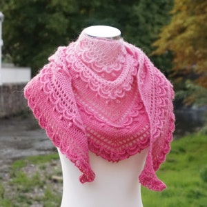 May include: A pink and white crocheted shawl with a delicate lace pattern. The shawl is draped over a white mannequin.