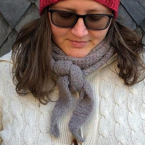 May include: A person wearing a red knit beanie, black sunglasses, a gray scarf, and a white cable-knit sweater. The background is a dark gray, diamond-patterned wall. The beanie is a solid, deep red color.