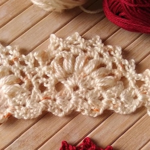 May include: Two crocheted lace trims, one in cream and one in red, are shown on a wooden surface. The trims are made with a delicate, intricate pattern.