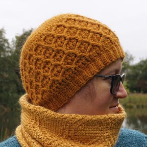 Puede incluir: Conjunto de gorro y cuello de punto amarillo mostaza. El gorro presenta un patrón de panal texturizado, y el cuello tiene un diseño acanalado. El conjunto está hecho de un hilo suave y cálido, perfecto para el clima frío. La persona lleva gafas de sol negras.