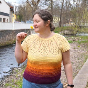 Puede incluir: Una blusa de crochet de manga corta con un degradado de color. La blusa pasa del amarillo en los hombros al naranja y luego al morado en la cintura. La blusa tiene un diseño calado detallado alrededor del escote.