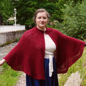 Może przedstawiać: Kobieta w czerwonym, dzianym szalu z koronkowym wzorem. Szal jest zarzucony na ramiona i ma falbankowy brzeg. Stoi przed strumieniem i zielenią.