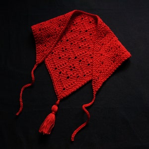 May include: A red crocheted cowl with a tassel. The cowl has a triangular shape and is tied with two long strings.