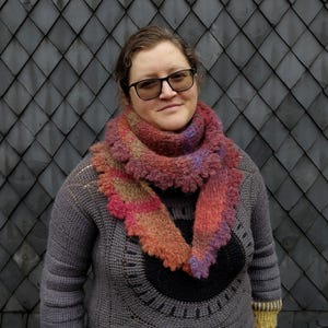 May include: A hand-knitted triangular scarf featuring a variegated color palette of orange, pink, purple, and brown tones. The scarf has a textured, fuzzy appearance with a ruffled edge and is worn wrapped around the neck over a gray crochet sweater.