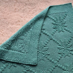 Könnte beinhalten: Eine türkisfarbene Strickdecke mit einem strukturierten Muster. Die Decke ist gefaltet und zeigt das komplizierte Design. Das Muster zeigt erhabene, stilisierte Blattformen, die in einem Rautenmuster angeordnet sind. Die Decke ist aus weichem Garn.