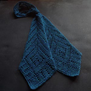 Puede incluir: Una bufanda de punto color verde azulado oscuro con un intrincado patrón de diamantes calados en toda la tela. La bufanda tiene una forma larga y cónica con un extremo en bucle, mostrando una textura detallada similar al encaje.