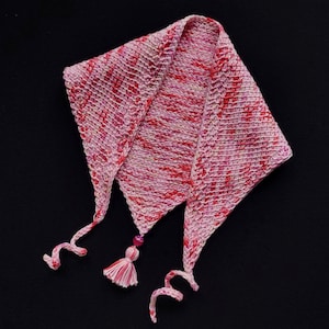 Op de afbeelding: Een roze en witte gehaakte sjaal met een kwast en een kraal. De sjaal is driehoekig van vorm en heeft een lang, vloeiend ontwerp.