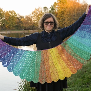 May include: A rainbow crochet shawl with a scalloped edge. The shawl is held by a person wearing a black dress and glasses.