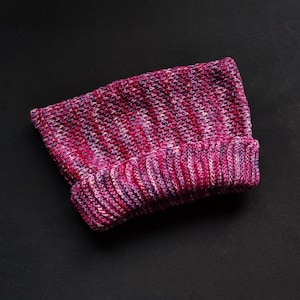 Puede incluir: Un gorro tejido en tonos de rosa, morado y blanco. El gorro tiene un borde doblado y un patrón de tejido texturizado. El gorro está hecho de un material suave y cálido.