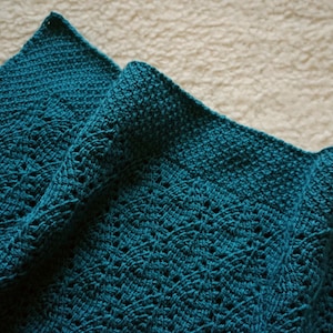 May include: A teal crocheted blanket with a lacy pattern. The blanket is folded in half and the edges are visible.