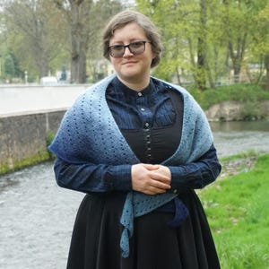 May include: A woman wearing a blue and black outfit, including a shawl with a gradient of blue hues. The shawl is draped over a dark blue plaid shirt and a black dress. She is wearing glasses and standing near a river.