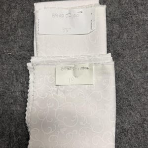 Könnte beinhalten: Zwei gefaltete weiße Stoffstücke mit einem zarten, wirbelnden Muster. Eines ist mit "89025-100" beschriftet und misst 83,8 cm. Das andere ist mit "89025-100" beschriftet und misst 25,4 cm. Der Stoff hat eine strukturierte Optik.