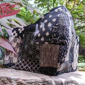 May include: A black and white patterned face mask made of fabric. The mask has a patchwork design with different geometric patterns.