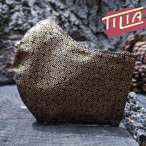 Könnte beinhalten: Schwarze Gesichtsmaske mit goldenem geometrischem Muster. Die Maske hat ein Tilia-Logo auf der rechten Seite.