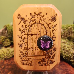 May include: A light-colored wooden plaque featuring a wood-burned illustration of an arched door surrounded by vines, flowers, mushrooms, a dragonfly, and bees. A circular black knob with a purple butterfly graphic is attached to the door.
