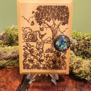 May include: A rectangular light-colored wooden plaque featuring a detailed pyrography illustration of a tree, flowers, and butterflies. A circular knob with a colorful hummingbird and floral graphic is attached to the right side of the wood.