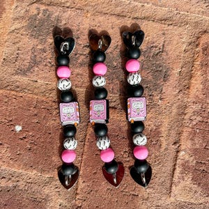 May include: Three beaded bookmarks with heart-shaped charms. Each bookmark features black, pink, and white beads, with a small book charm that reads "Bookish Girlie". The bookmarks have a heart-shaped charm at the top and bottom.