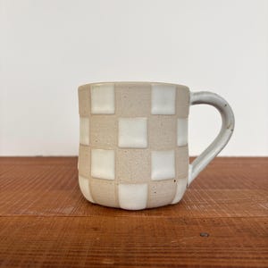 May include: A ceramic mug with a checkerboard pattern of beige and white squares. The mug has a curved handle and sits on a wooden surface. The mug is a neutral color and would be suitable for coffee or tea.