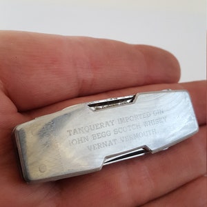 Op de afbeelding: Een zilveren multitool met een vouwmes en een verscheidenheid aan andere gereedschappen. De tool is gegraveerd met de tekst "Tanqueray Imported Gin, John Begg Scotch Whisky, Vernat Vermouth."