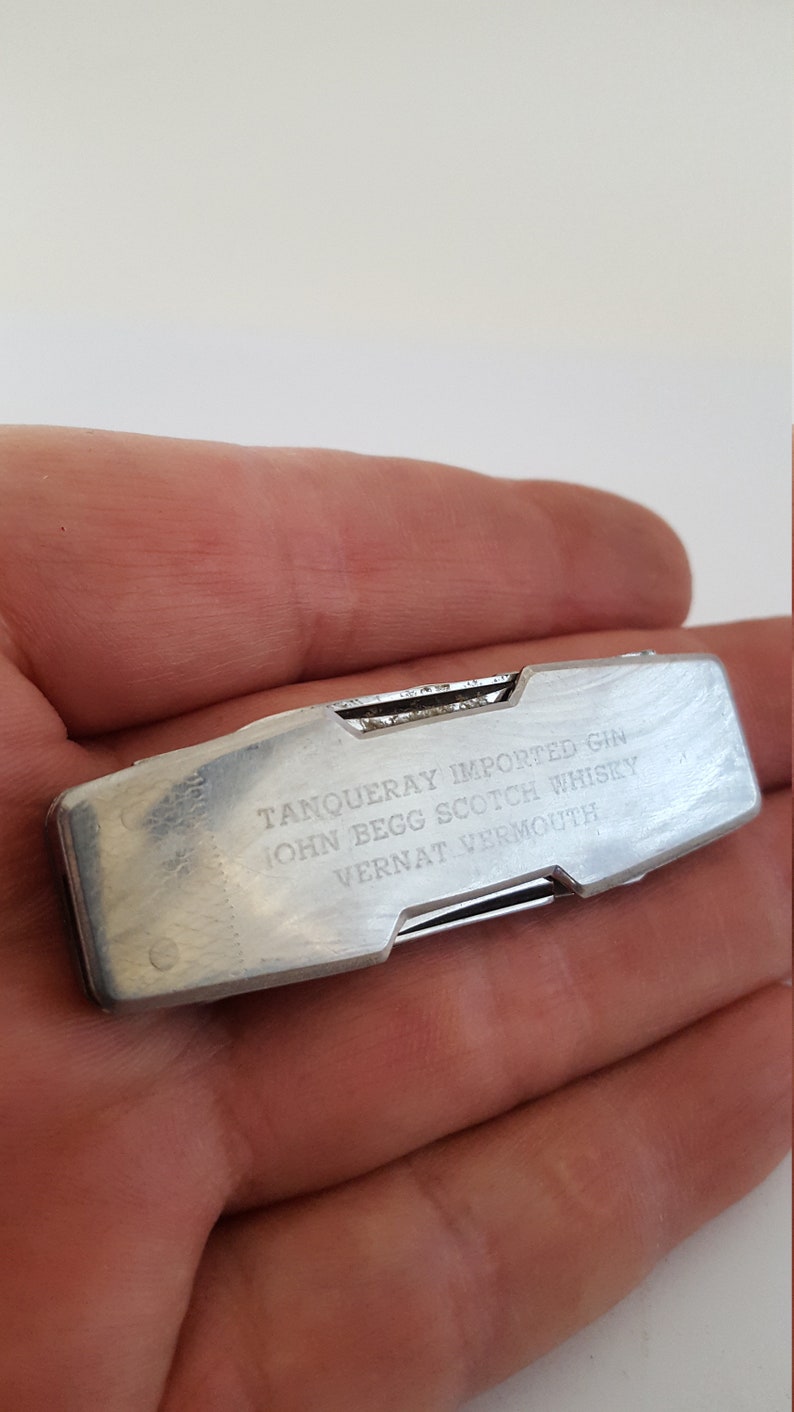 Op de afbeelding: Een zilveren multitool met een vouwmes en een verscheidenheid aan andere gereedschappen. De tool is gegraveerd met de tekst "Tanqueray Imported Gin, John Begg Scotch Whisky, Vernat Vermouth."