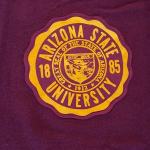 May include: A maroon patch with a yellow border and the text "Arizona State University" in a circular design. The patch also features a smaller seal with the text "Great Seal of the State of Arizona" and "Ditat Deus" with a star on either side of the year "1912". The numbers "18" and "85" are on either side of the text "Arizona State University".