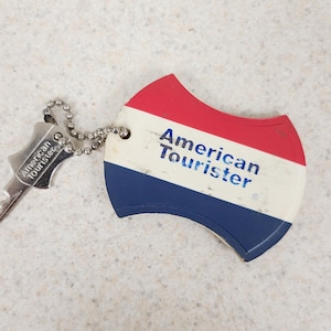 May include: A metal key and a curved, shield-shaped plastic luggage tag connected by a silver ball chain. The tag features horizontal red, white, and blue stripes with the text American Tourister printed in blue on the white section.