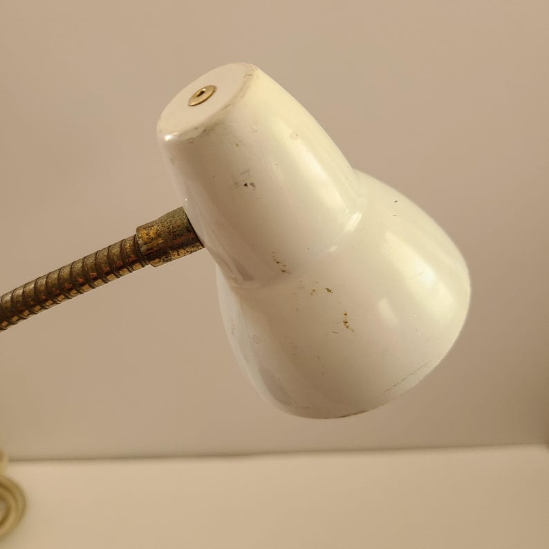 May include: A vintage white desk lamp with a flexible, brass-colored gooseneck. The lamp head is a rounded, cone shape with a small, circular opening on top. The lamp shows signs of wear.