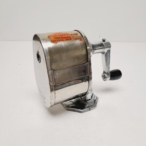 May include: A vintage silver-toned pencil sharpener with a black handle. The sharpener has a rectangular body with a crank and a base. The label reads "SKANS-SHEPPARD CO. 1316 Douglas St. OMAHA, NEBRASKA."