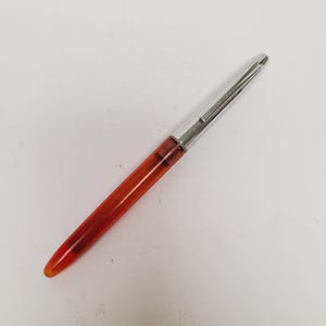 May include: A vintage fountain pen with a transparent orange barrel and a silver-colored cap. The pen is angled, showcasing its sleek design and the contrast between the vibrant barrel and the metallic cap. The pen is a writing instrument.