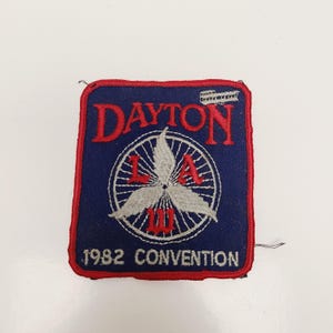 May include: A square patch with a blue background and red border. The word "DAYTON" is in red at the top. A white propeller design is in the center with the letters "L A III" in red. The text "1982 CONVENTION" is at the bottom.