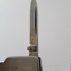 Op de afbeelding: Een zilveren multitool met een mes waarop "STAINLESS JAPAN" staat. De tool heeft een zwarte handgreep met de tekst "WED GIN WHISKEY MOUTH" erop.
