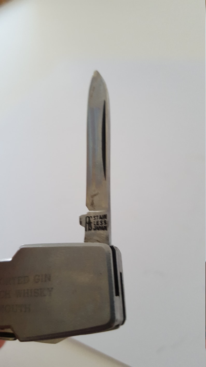 Op de afbeelding: Een zilveren multitool met een mes waarop "STAINLESS JAPAN" staat. De tool heeft een zwarte handgreep met de tekst "WED GIN WHISKEY MOUTH" erop.