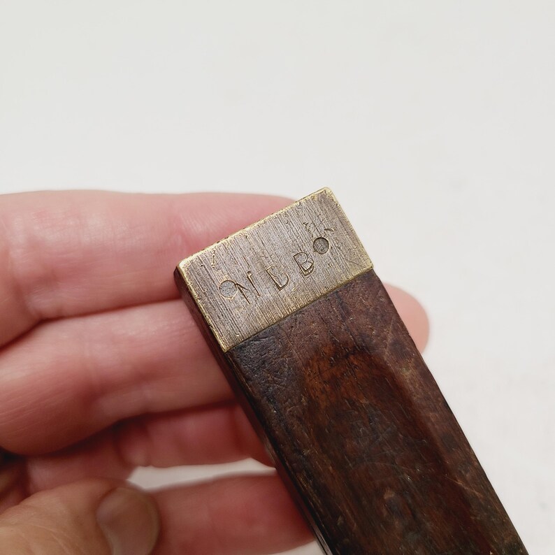 May include: A rectangular wooden handle featuring a brass end cap. The brass cap is engraved with the letters NLBO. The wood has a dark brown, textured grain, and the metal component shows a brushed, metallic finish with visible surface markings.