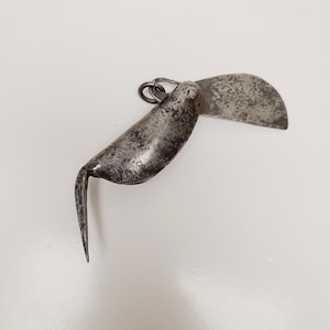 May include: An antique, silver-toned folding knife with a curved blade and a pointed hook. The blade is open, revealing a textured surface. The knife has a ring for attachment and shows signs of wear, suggesting age and use.