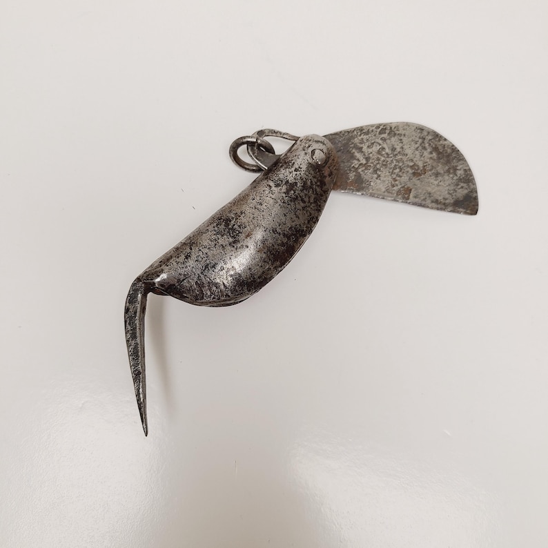 May include: An antique, silver-toned folding knife with a curved blade and a pointed hook. The blade is open, revealing a textured surface. The knife has a ring for attachment and shows signs of wear, suggesting age and use.