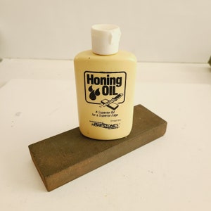 May include: A yellow bottle of honing oil with a white cap. The bottle has black text that reads "Honing Oil" and "A Superior Oil for a Superior Edge". The bottle also has a black image of a knife and three drops of oil. The bottle is sitting on a brown sharpening stone.
