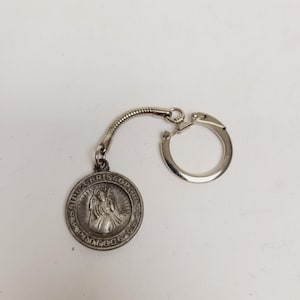May include: Silver-toned Saint Christopher keychain. The round pendant features an image of Saint Christopher and the words "SAINT CHRISTOPHER PROTECT US". The keychain has a coiled connector and a split ring for keys.