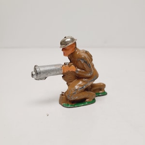May include: A vintage toy soldier figurine, kneeling and aiming a silver machine gun. The soldier wears a brown uniform and a silver helmet. The base is green. The toy has visible signs of wear.