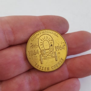 May include: A gold-colored coin commemorating the Lassen County Centennial. The coin features a wagon wheel with a sunburst design and the years 1864 and 1964. The text "Centennial" and "Lassen County" are inscribed around the edge.