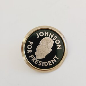 May include: A gold-colored button with a black background and white text. The text reads "JOHNSON FOR PRESIDENT". A profile image of a man is in the center of the button.