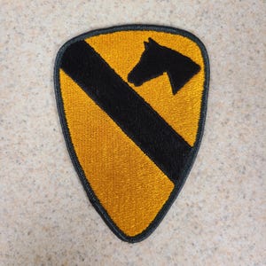 May include: A shield-shaped embroidered patch featuring a golden yellow background with a thick black diagonal stripe running from the top left to the bottom right and a black horse head silhouette positioned in the upper right section.