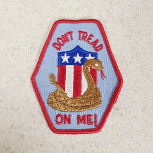 May include: A hexagonal embroidered patch with a light blue background and a red border. It features a brown coiled snake in front of a shield with red and white vertical stripes and three white stars on a blue field, with the text DONT TREAD ON ME.