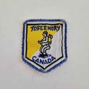 May include: A blue and yellow fabric patch with a cartoon figure of a person wearing a backpack and the text "TOBERMORY CANADA".
