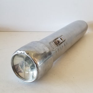 May include: A silver flashlight with a black switch and the word "DELUXE" printed on the side. The flashlight has a clear glass lens.