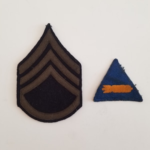 May include: Two vintage military patches. One is a dark blue chevron with three brown stripes, and the other is a blue triangle with an orange design. Both patches are on a white background.