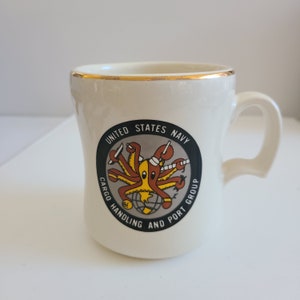 May include: White ceramic mug with a gold rim and a black and white logo featuring an octopus with a sailor hat and the text "United States Navy Cargo Handling and Port Group".