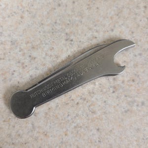 Op de afbeelding: Een zilveren metalen flesopener met de tekst "HUTCHINSON METAL DECORATING COMPANY BIRMINGHAM, ALABAMA" gegraveerd op het handvat.