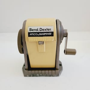 May include: A vintage Berol Dexter pencil sharpener with a yellow plastic body and a gray metal base. The sharpener has a crank handle and a label that reads "Berol Dexter APSCO SHARPENER".