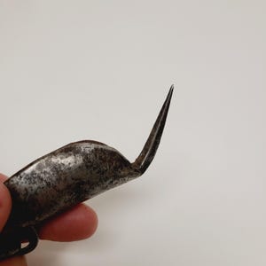 May include: Close-up of a vintage, curved metal tool with a sharp, pointed end. The tool has a worn, aged appearance, with a curved handle and a pointed blade. The metal has a dark, weathered finish, suggesting age and use.